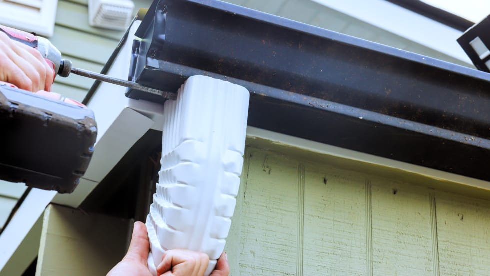 A contractor using a power drill to attach a white downspout elbow to a dark eavestrough along the roofline of a house.