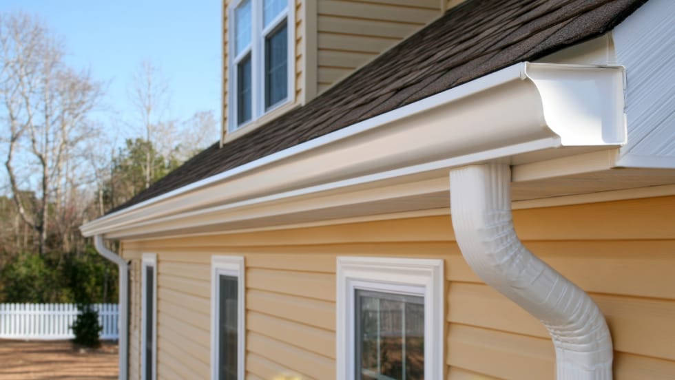 A clean, white seamless eavestrough system with an attached downspout running along the roofline of a home with yellow siding.